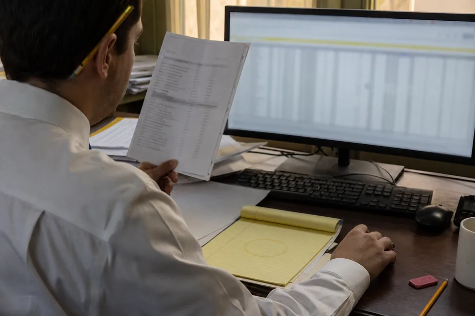 Staff accountant comparing a printed bank statement with a spreadsheet on a monitor